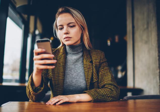 A young woman with a serious expression on her face is sitting at a table, using a smartphone. Image by GaudiLab via Shutterstock.