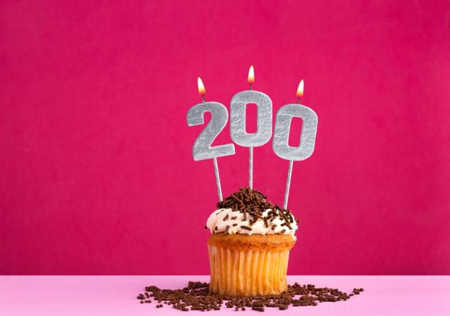 A birthday cupcake with lit silver candles saying '200'. The cupcake has white icing and chocolate sprinkles. The background of the image is pink. Image by Luis Echeverri Urrea via Shutterstock.