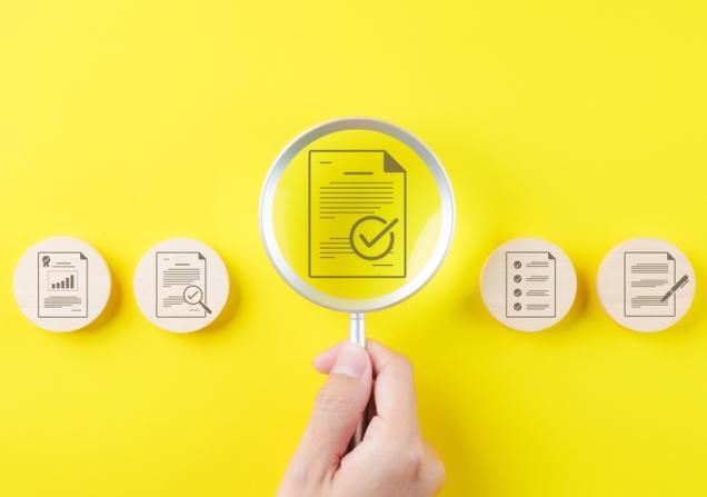 In the centre of the image, a hand is holding a magnifying glass over an icon of a document with a tick mark on it. On either side are more icons with documents and tick marks on wooden circles. The background of the image is bright yellow. Image by SuPatMaN via Shutterstock.