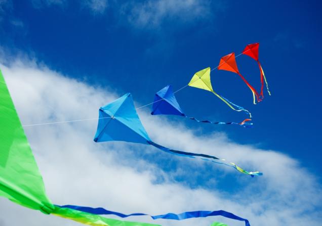 A string of coloured kites flying high against a blue sky with a few white clouds. Image by Sergey Novikov via Shutterstock.