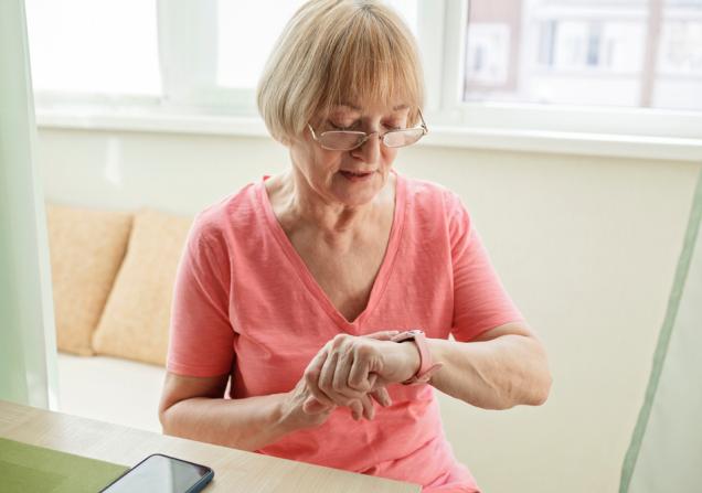 A senior woman is looking at a fitness tracker on her wrist. She has short blonde hair and glasses and is wearing a pink t-shirt. Image by Maria Symchych via Shutterstock.